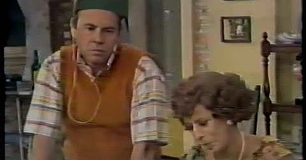 A man in a striped shirt and orange vest sits at a table, looking concerned, with a woman in a floral dress beside him.