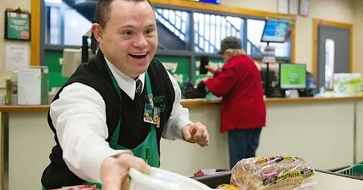 A smiling man in a green vest hands out a bag of groceries in a store.