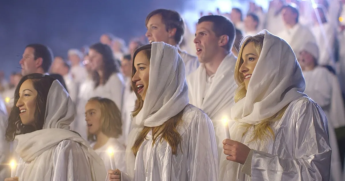 A group of people in white robes and head coverings hold candles, smiling and looking forward.