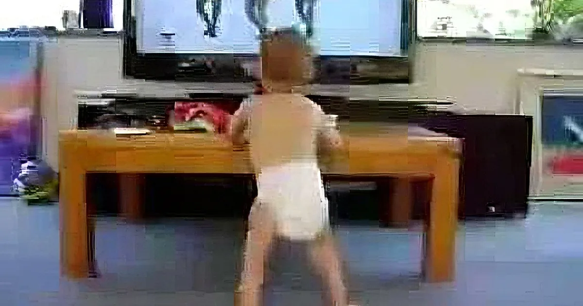 A toddler in a diaper stands on a chair, watching TV, with a colorful room and furniture in the background.