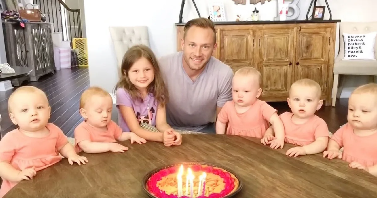 A man and six children gather around a table with a cake, celebrating a special occasion.