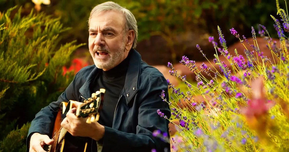 A man in a dark jacket plays a guitar amidst vibrant purple flowers and greenery.