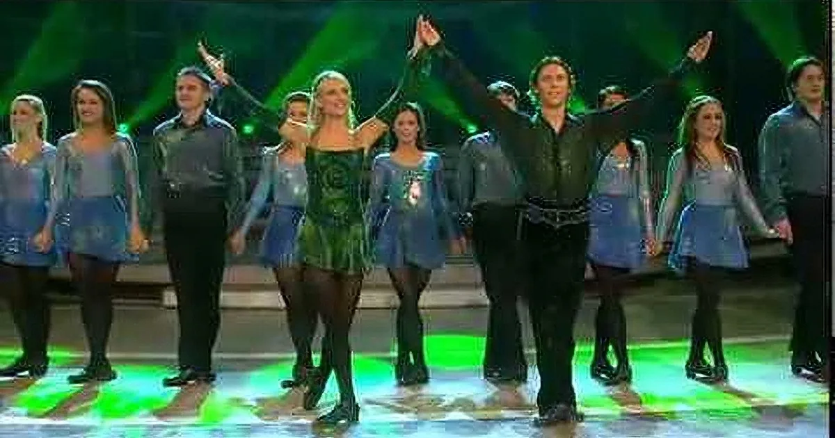 A person in a black outfit stands on a stage with green lights and a reflective floor.