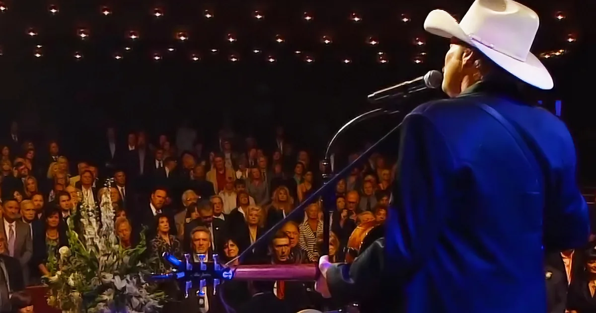 A man in a white cowboy hat sings into a microphone on stage in front of a large audience.
