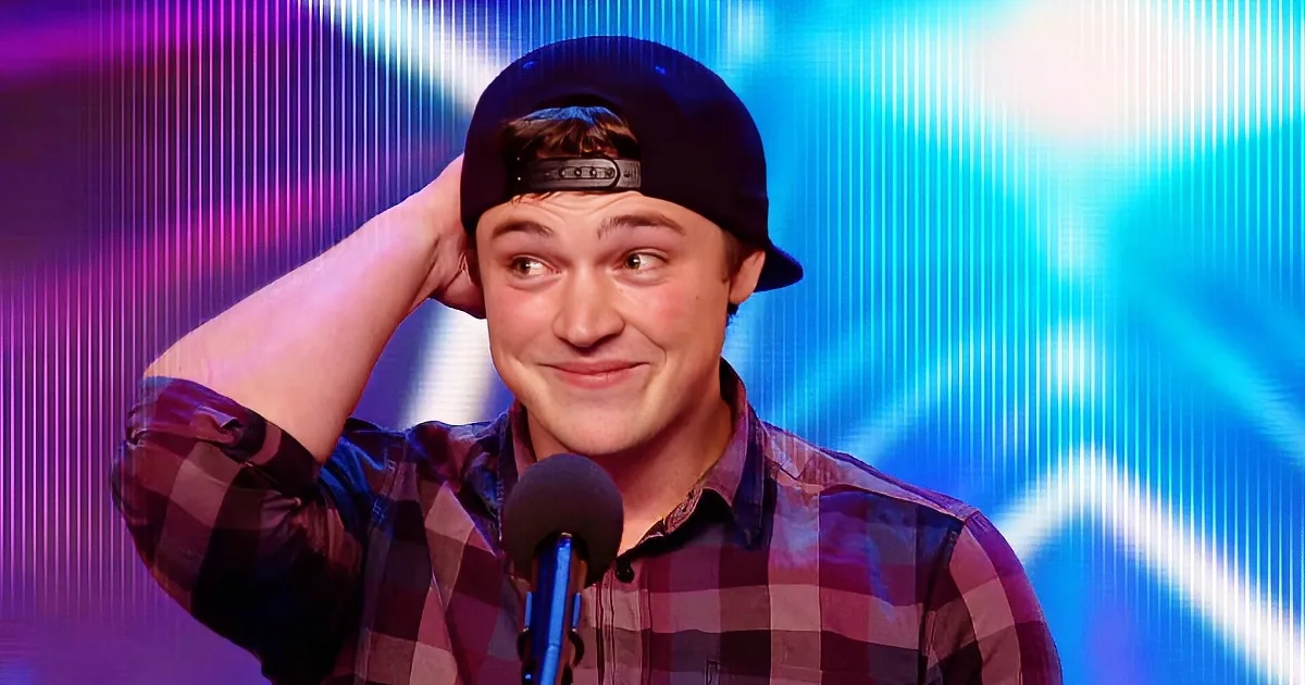 A person in a plaid shirt and cap is holding their head, smiling, with a microphone in front of them against a colorful...