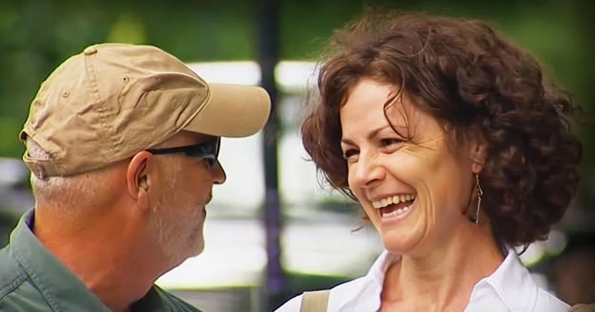 A man in a cap and sunglasses smiles at a woman with curly hair, both laughing.