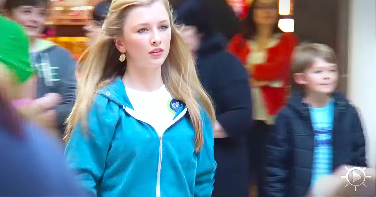A young woman with long blonde hair wears a teal jacket and white shirt, standing in a crowd.