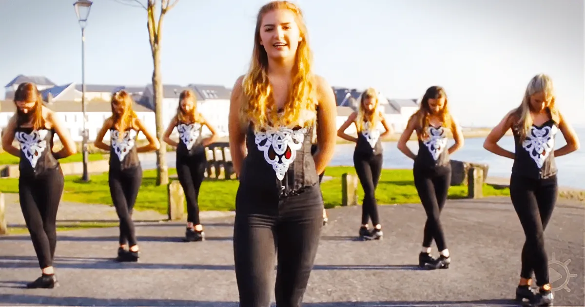A woman in a black outfit with white corset stands in front of a group of dancers in matching outfits.