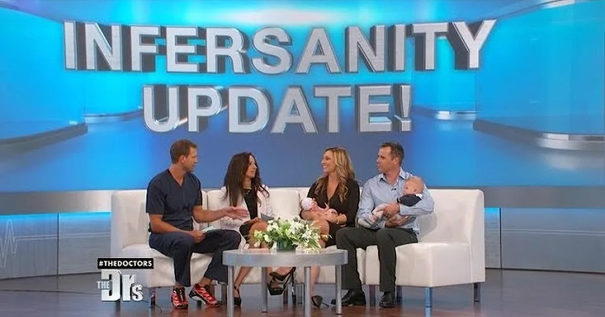 A man in a light blue shirt holds a baby while smiling at a woman in a black top on a talk show set.