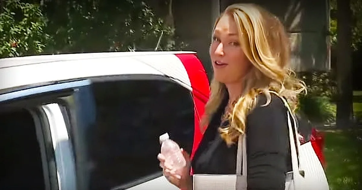 Blonde woman in car, holding water bottle, seatbelt on, trees and car visible outside.