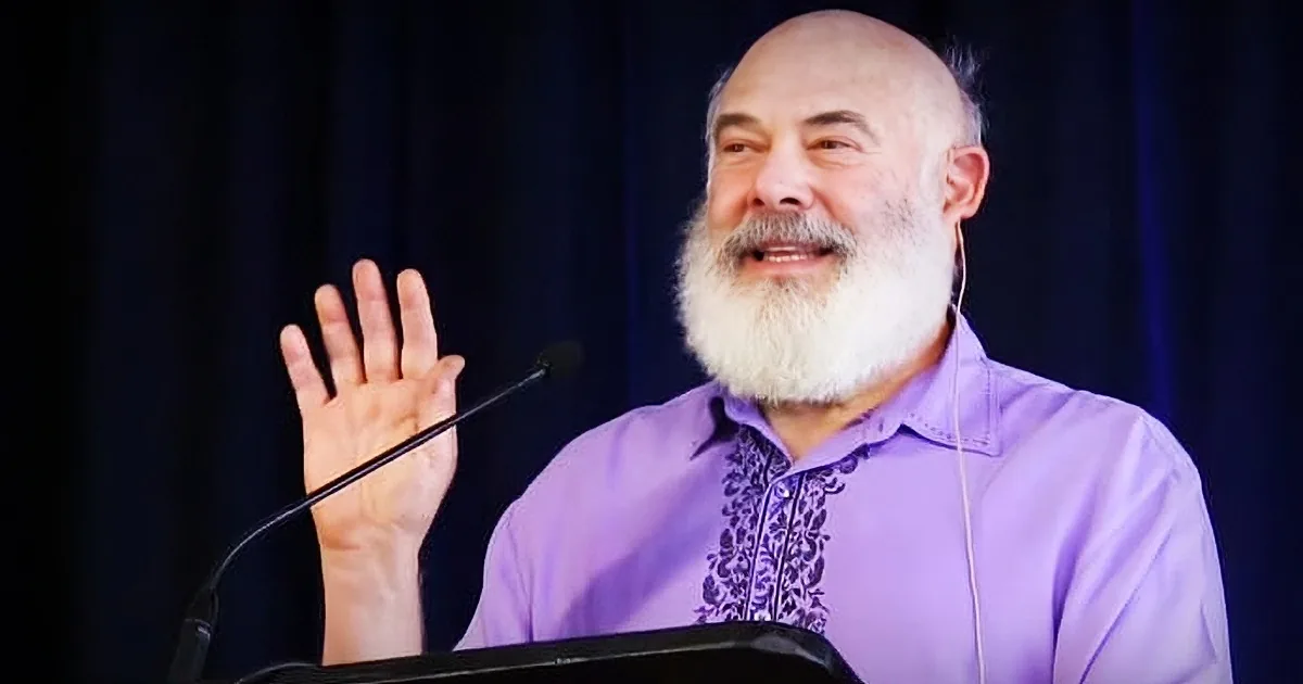 A man with a white beard and purple shirt is speaking into a microphone, gesturing with his hand.