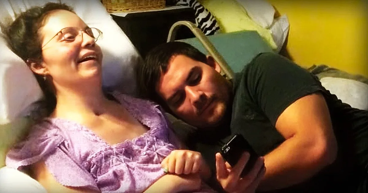A woman in a hospital bed, smiling, with a man lying beside her, holding her hand.