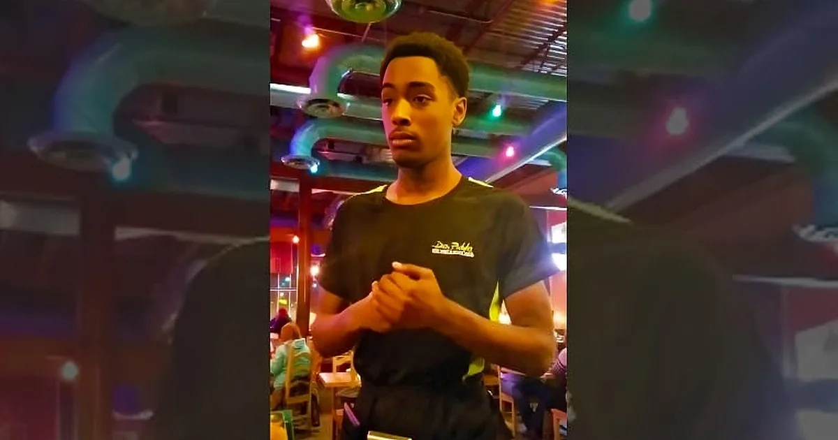 A young man in a black shirt stands in a colorful, dimly lit room with a ceiling fan and lights above.