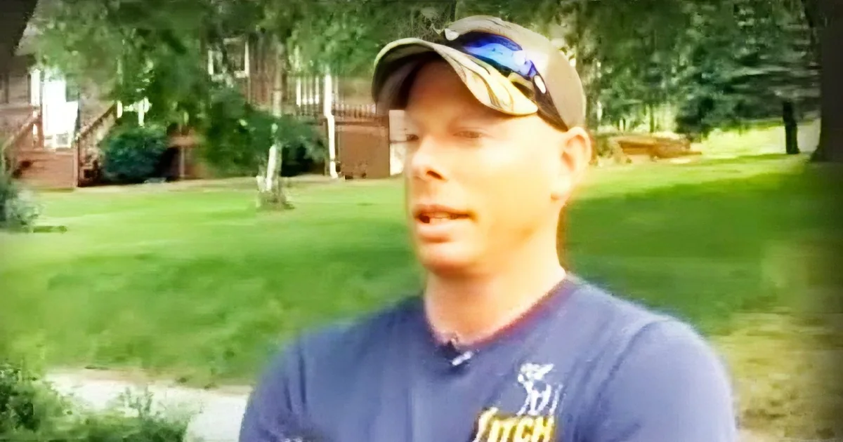 A man wearing a blue shirt and cap stands outdoors, holding a book, with a grassy yard and trees in the background.