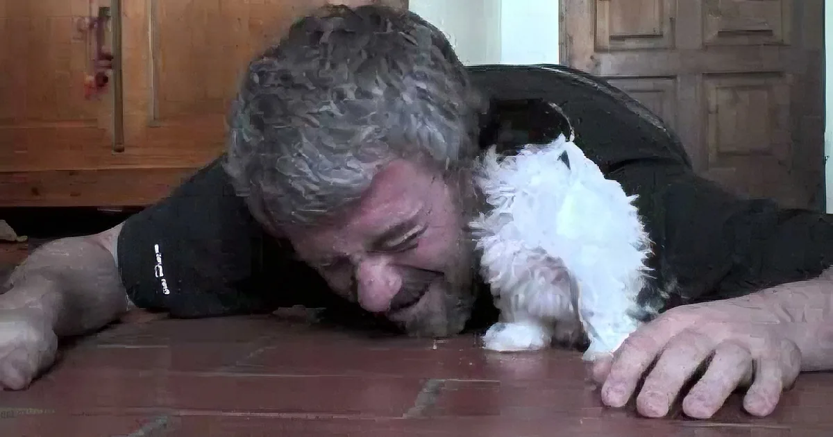 A man lies on a wooden floor, embracing a small white and black dog closely.