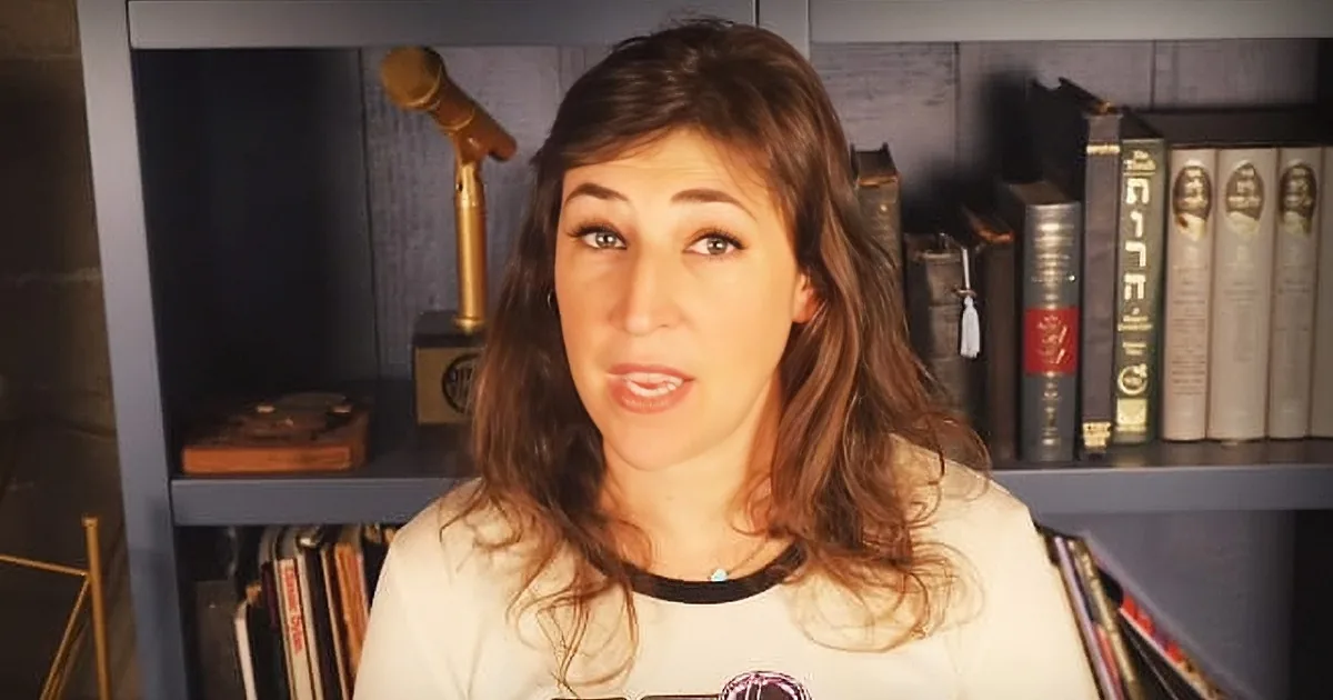 A woman with long brown hair and a white shirt stands in front of a bookshelf with books and a Darth Vader poster.