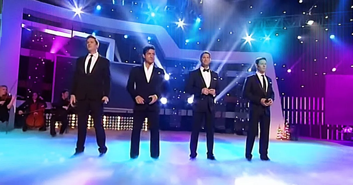 Four men in tuxedos stand on a stage with colorful lights and a band in the background.