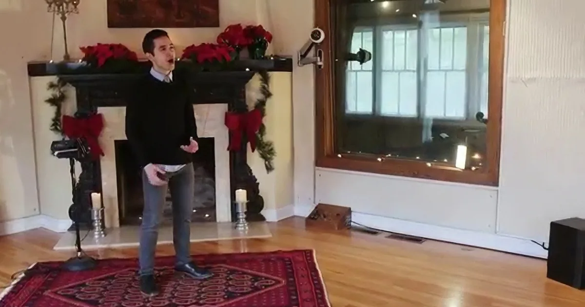 A man stands on a red rug in front of a decorated fireplace, singing with a microphone.