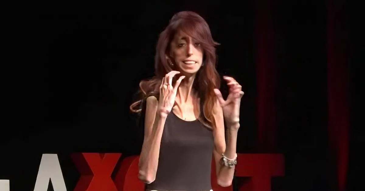 A woman with long brown hair gestures with her hands while speaking on stage at a TEDx event.