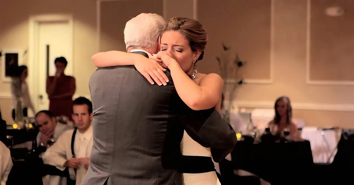 A woman in a white dress hugs a man in a gray suit at a formal event.