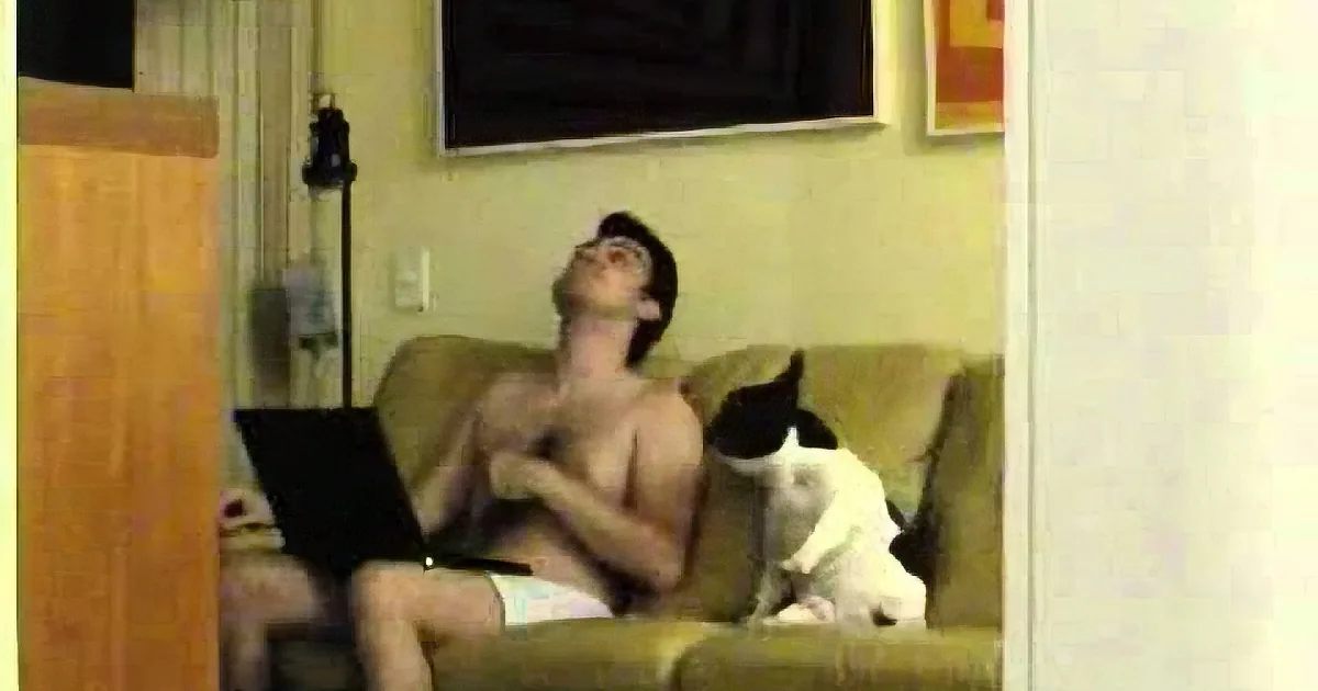 A shirtless man sits on a couch, holding a laptop, with a black and white cat beside him.
