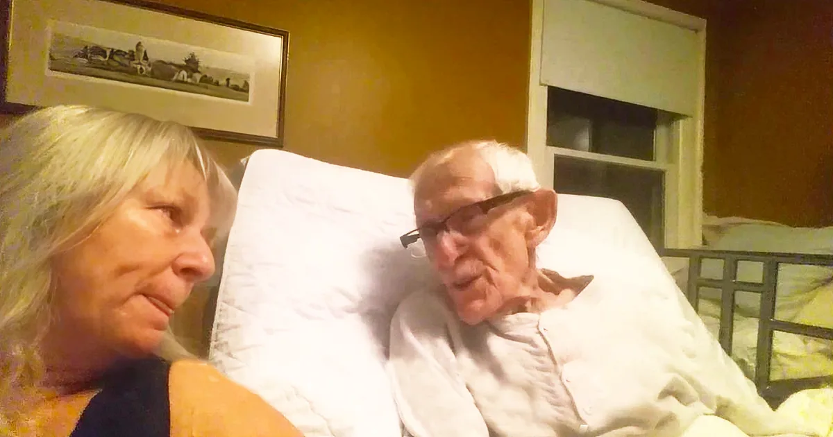 An elderly man in a hospital bed, wearing glasses, with a woman sitting beside him.