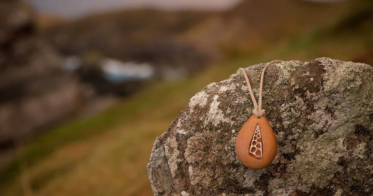 A teardrop-shaped pendant with a geometric cut-out hangs from a string on a mossy rock.