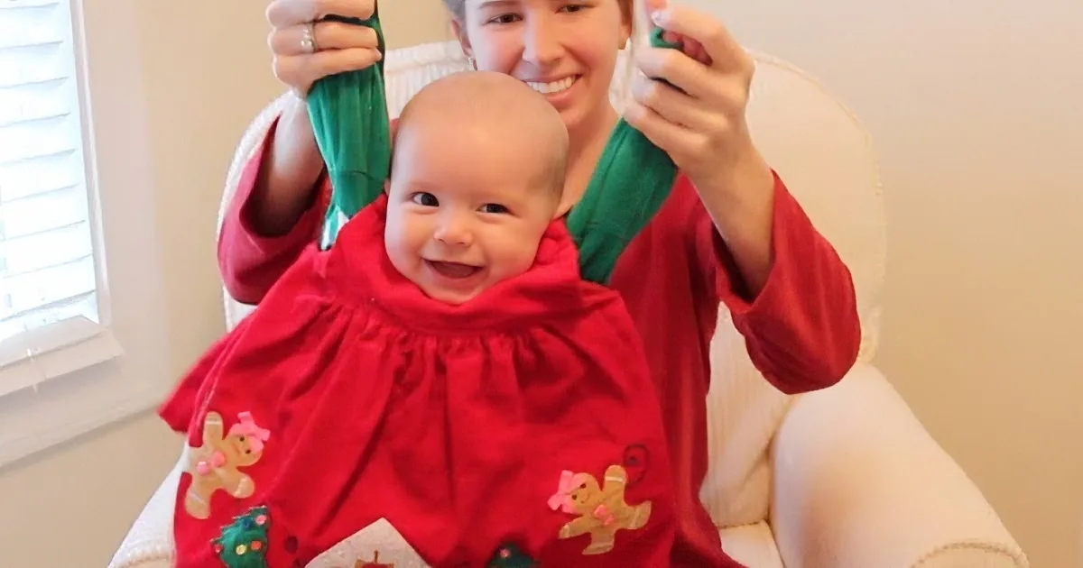 A baby in a red Christmas dress smiles while being held by a person in a red shirt.