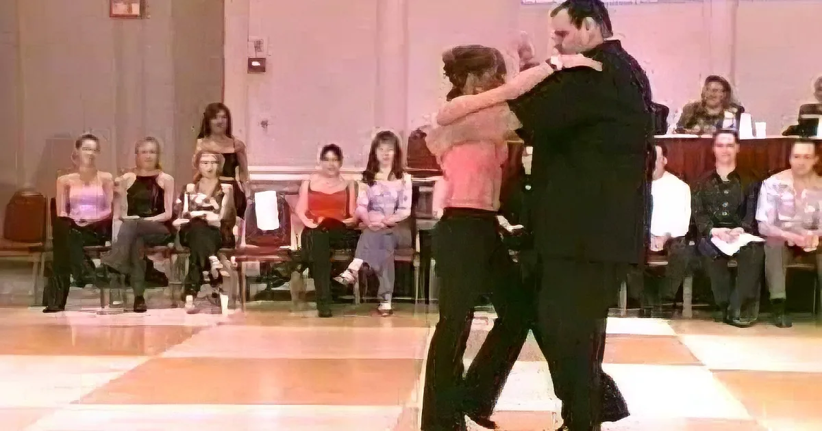 A couple dances in a ballroom with spectators watching in the background.