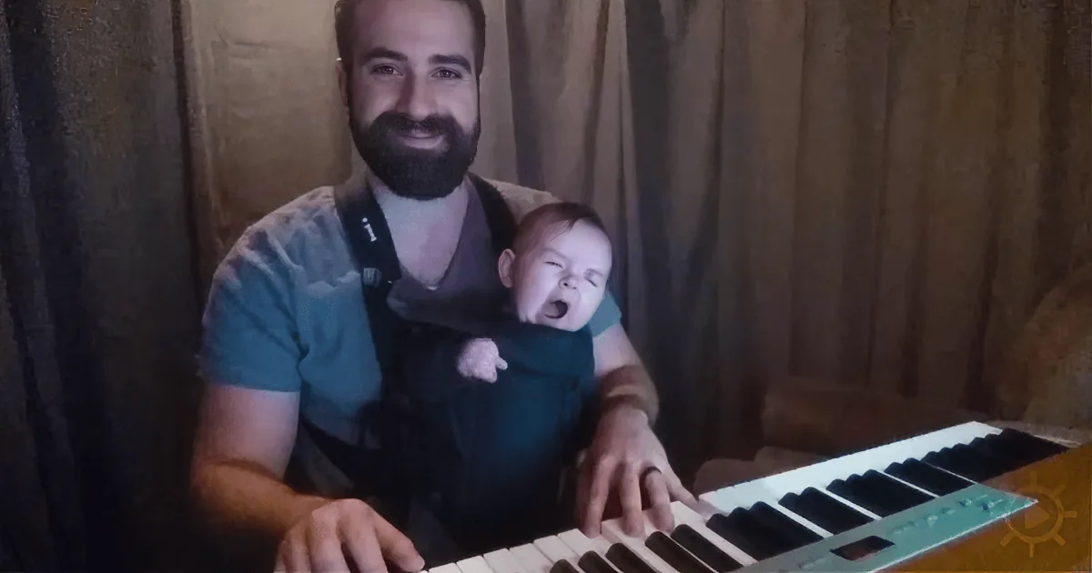 A man with a beard holds a baby in a carrier while playing a keyboard.