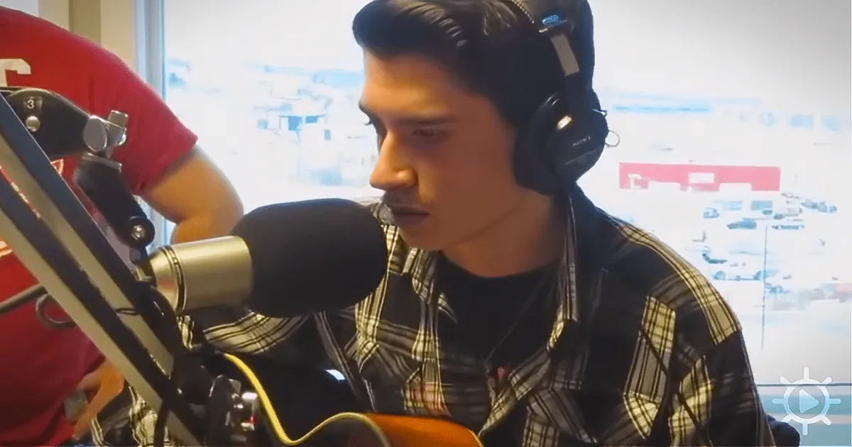 A person wearing headphones and a plaid shirt is singing into a microphone while holding a guitar.