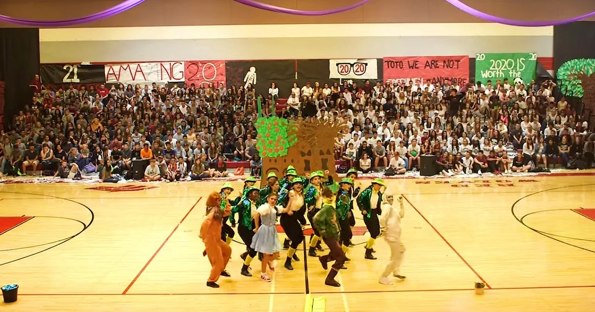 Students in costumes perform on a gymnasium floor with a large audience and banners in the background.