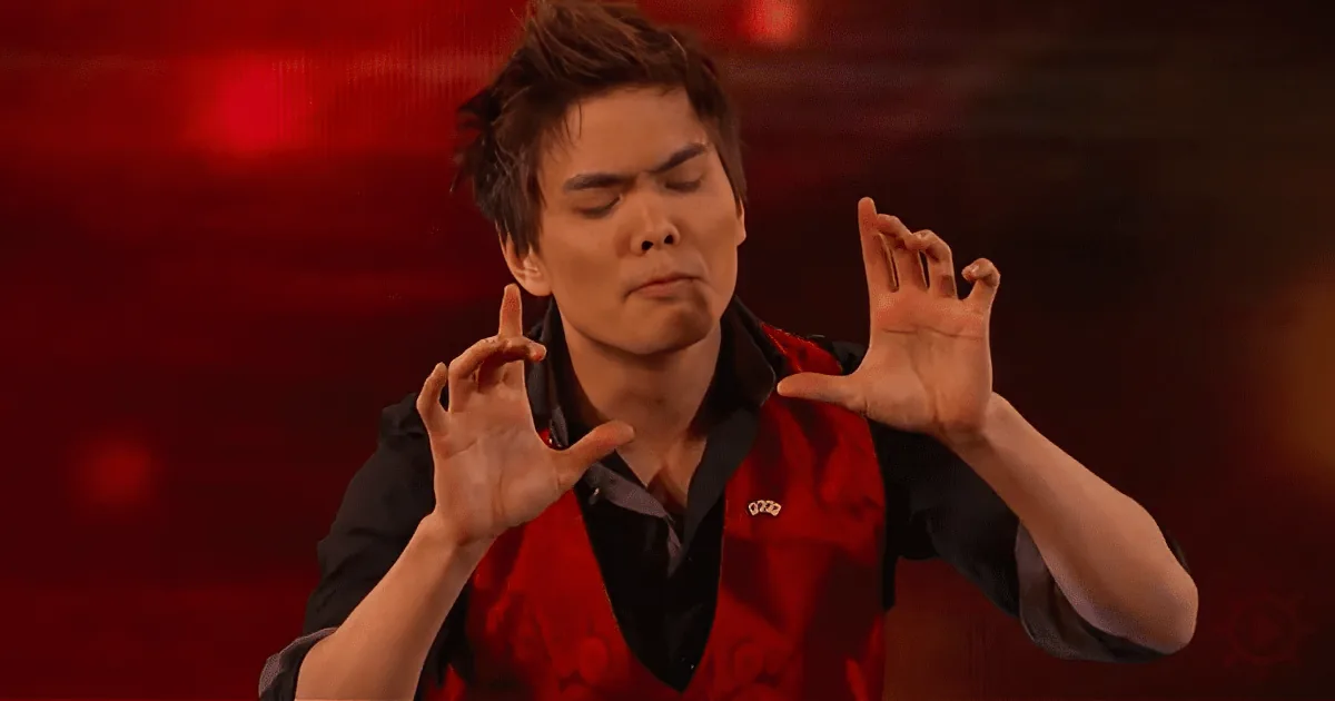 A person in a red vest and black shirt makes a gesture with their hands against a red background.