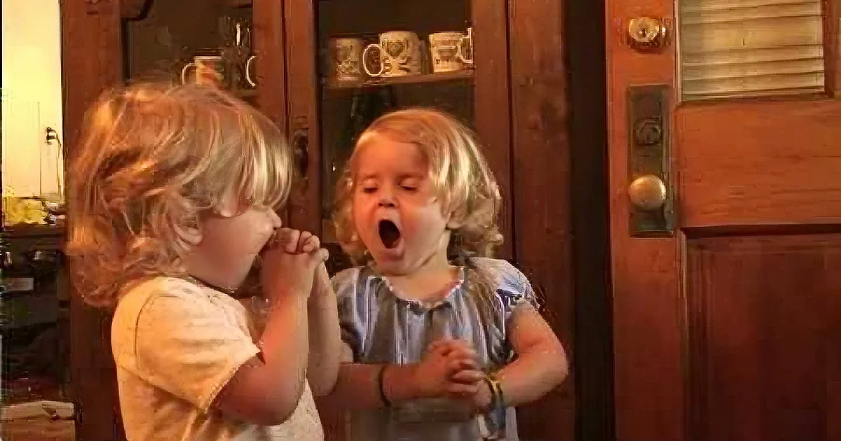Two children, one with hands over mouth, the other with mouth open, standing in front of a wooden door with a glass panel.