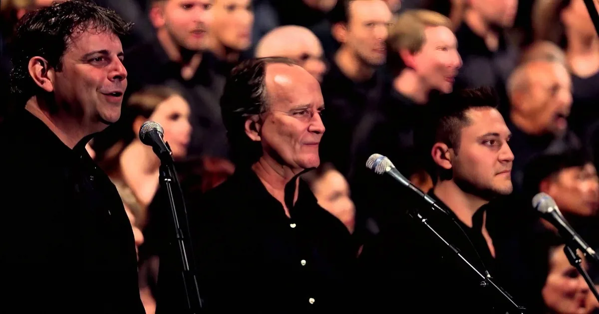 A man sings into a microphone, surrounded by a choir in black shirts, with a blurred audience in the background.