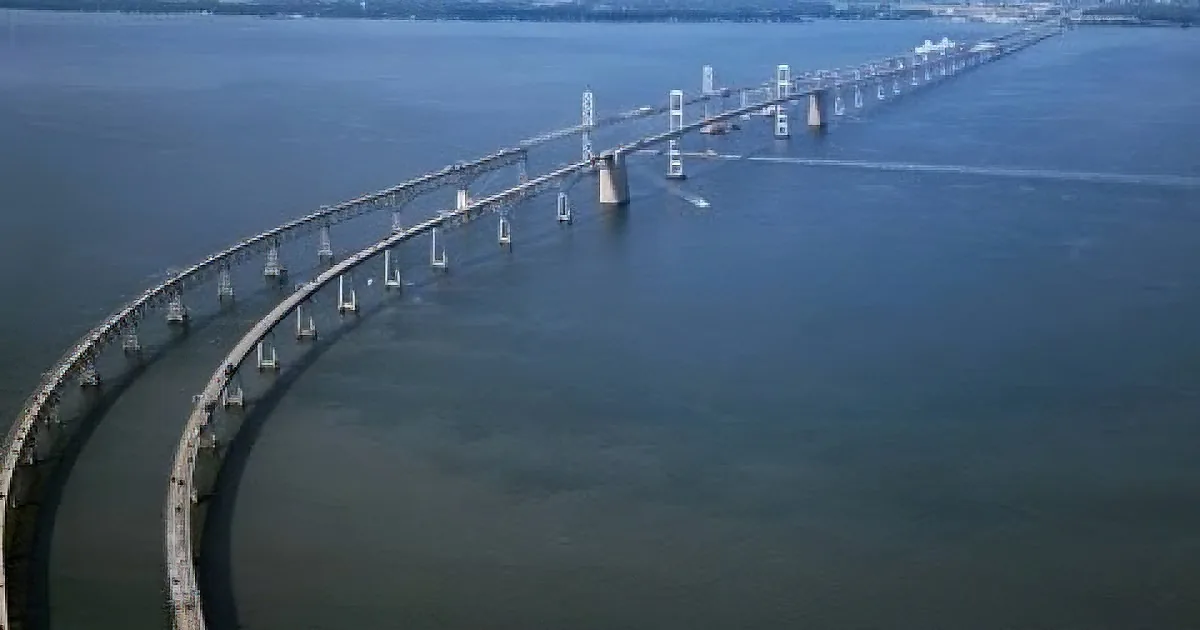A long, curved bridge spans a large body of water, with a boat visible in the distance.