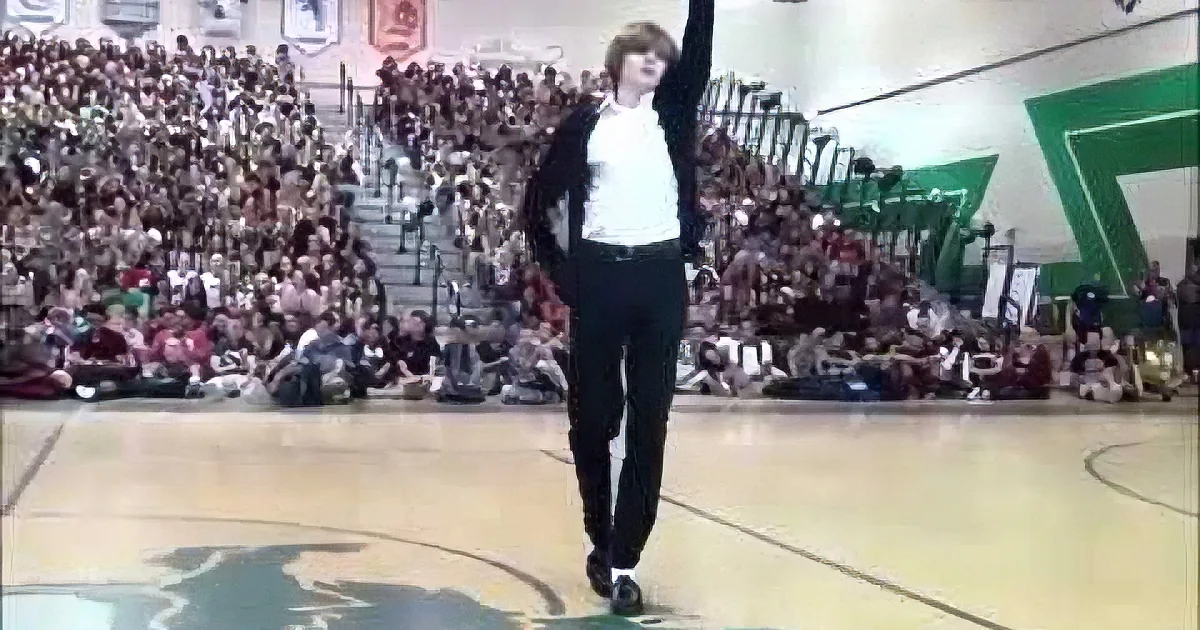 A person in a white shirt and black pants raises one arm, standing on a basketball court in front of a large audience.