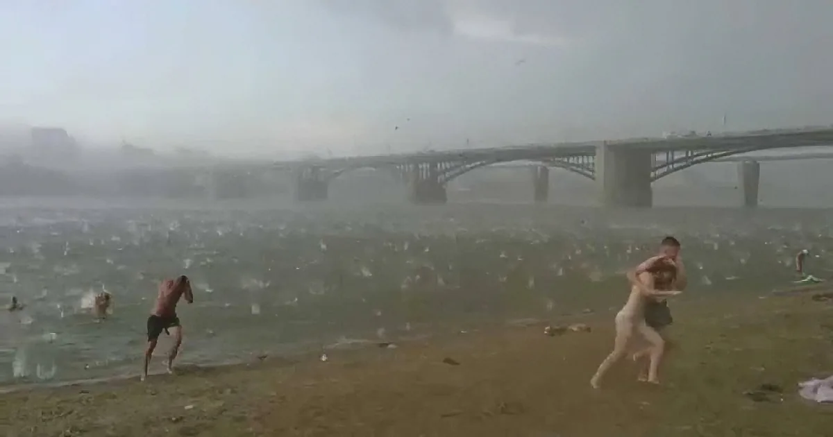 Two shirtless men wrestle on a beach as strong winds blow debris and water, with a bridge in the background.
