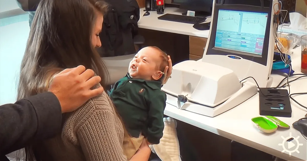 A woman holds a baby in front of a computer monitor displaying a graph.