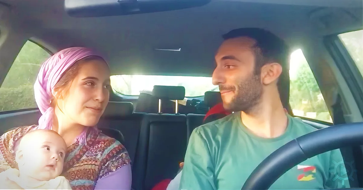 A family of three is in a car, with the man driving and the woman holding a baby, all smiling.