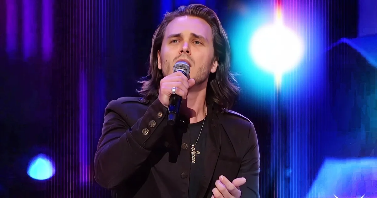 A man with long hair and a beard sings into a microphone on stage, wearing a black jacket and a cross necklace.