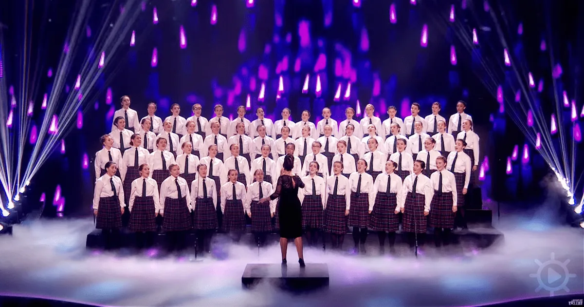 A conductor leads a choir in a formal performance with purple lighting and fog.