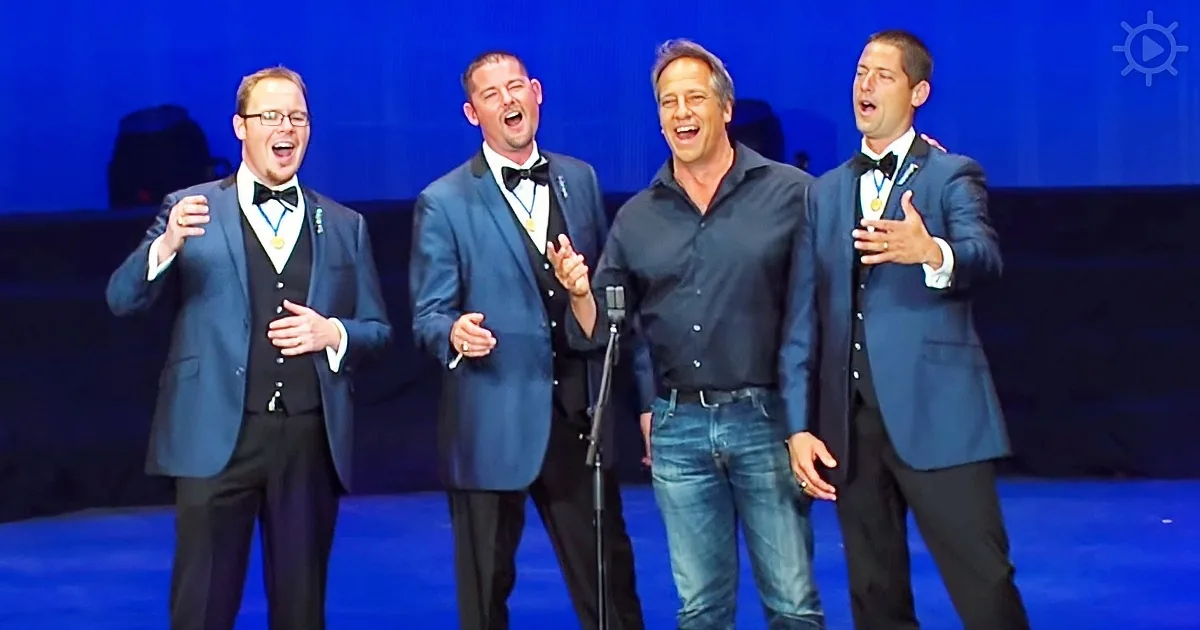 Four men in formal attire sing on stage, one in jeans, others in suits, with medals around their necks.