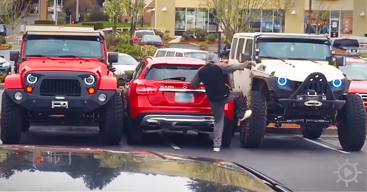 A man in a hoodie leans between two parked Jeeps in a parking lot.