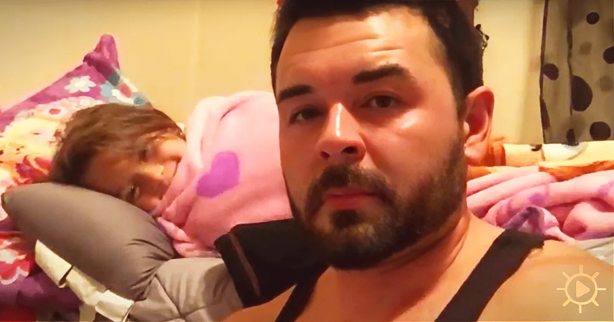 A man with a beard and a woman in the background, both in bed with colorful pillows and blankets.