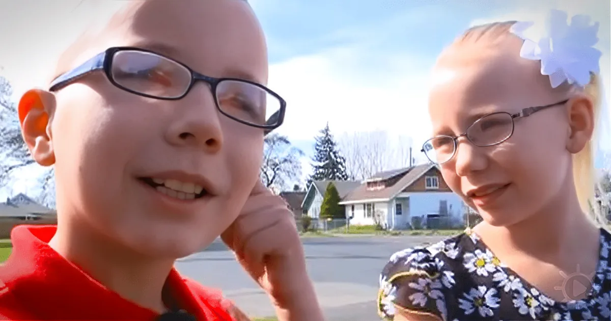 A boy in a red shirt and glasses smiles, with a girl in a floral dress and glasses beside him, both outdoors on a sunny day.