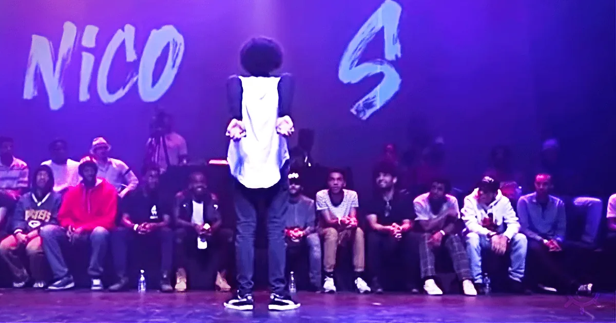 A person stands on stage with arms outstretched, surrounded by seated audience members under purple lighting.