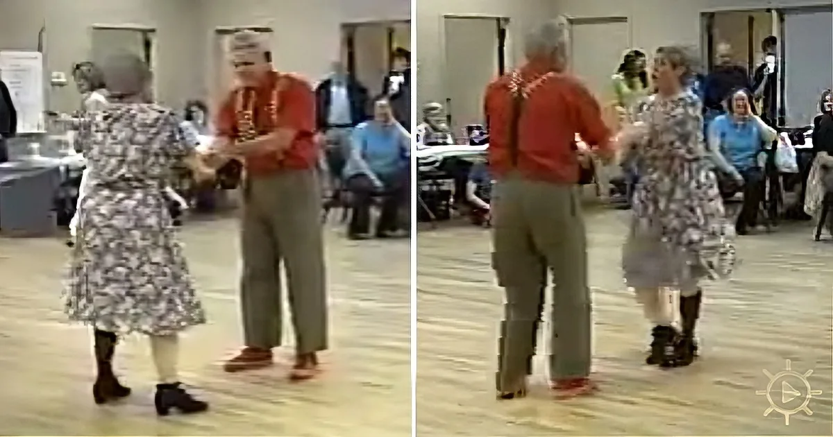 An elderly couple dances together in a community center, surrounded by other people.