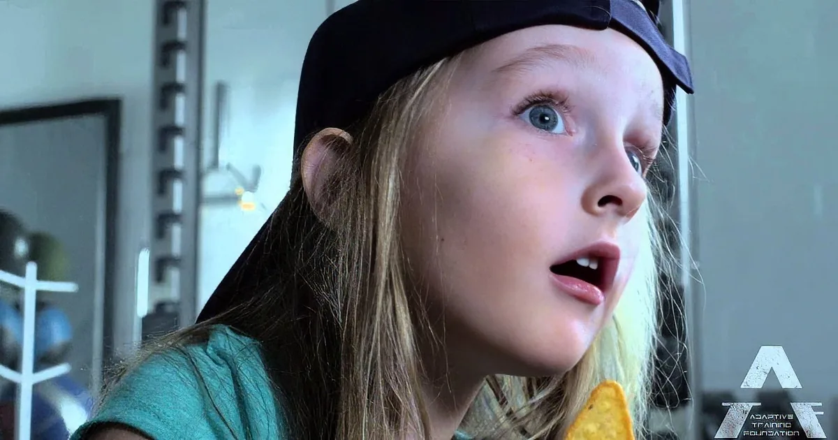 A young girl with long blonde hair and a black cap looks surprised, wearing a teal shirt.