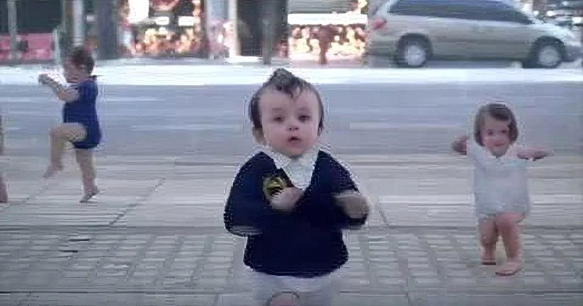 A baby in a Batman shirt stands on a sidewalk, surrounded by blurred images of other children playing.